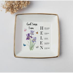 May include: A square, white ceramic trinket dish with a gold rim. The dish features the text "God Says I am" and the name "HELEN" spelled out vertically with inspirational words. Purple irises and butterflies decorate the dish.