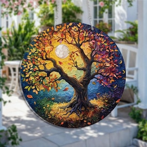 May include: A round, colorful glass suncatcher featuring a stylized tree with a full moon. The tree's leaves are in shades of red, orange, and yellow, with a dark brown trunk and branches. The background transitions from blue to yellow, with a crescent moon.