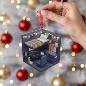 May include: A blue and white Christmas ornament shaped like a truck cabin interior. The ornament features a bed with a plaid blanket, a steering wheel, and a small shelf with the name "Henry" on it. A red ribbon is attached for hanging.