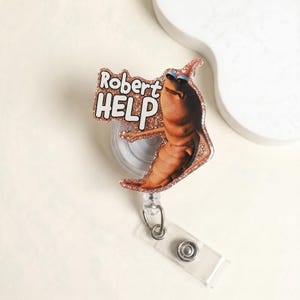 May include: A novelty retractable badge reel featuring a cartoonish, brown creature wearing sunglasses. The badge reel has a glittery rose gold border and the words "Robert HELP" in white.