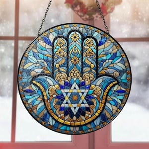 May include: Round stained glass window art featuring a Hamsa hand design. The artwork uses shades of blue, gold, and yellow in a mosaic pattern. A Star of David is at the center. The window is framed in black and hangs from a chain.