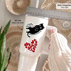 May include: A white tumbler with a handle, featuring stickers. One sticker has a black cat design and the text "I can do all things through shit which strengthens me." Another sticker says "You are loved!" in red. A ruler is in the background.