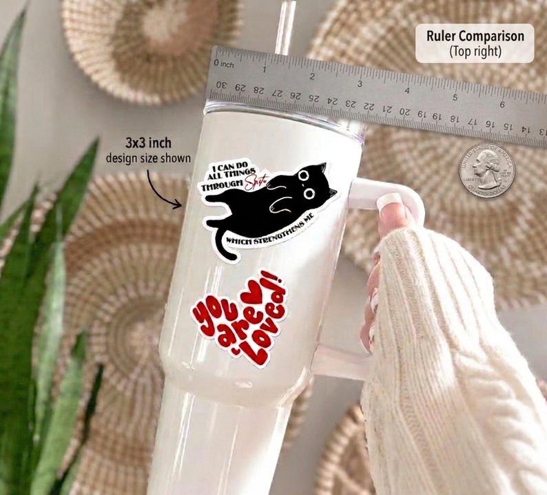 May include: A white tumbler with a handle, featuring stickers. One sticker has a black cat design and the text "I can do all things through shit which strengthens me." Another sticker says "You are loved!" in red. A ruler is in the background.