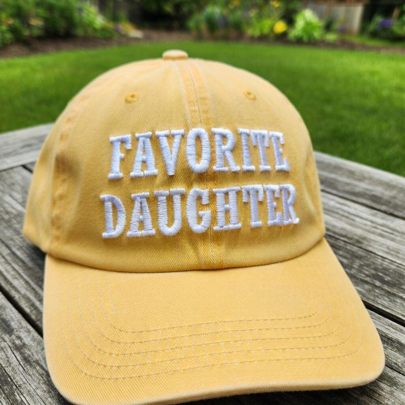 May include: A yellow baseball cap with the embroidered text "FAVORITE DAUGHTER" in white. The cap has a curved brim and a fabric strap closure. The background shows a wooden surface and green foliage.