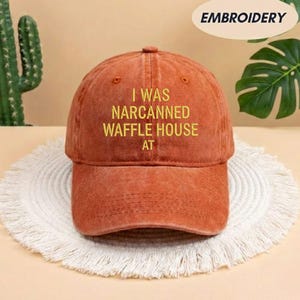 May include: A rust-colored baseball cap with the embroidered text "I WAS NARCANNED WAFFLE HOUSE AT" in yellow. The cap is sitting on a white, fringed placemat. The word "EMBROIDERY" is in a white oval in the upper right corner.