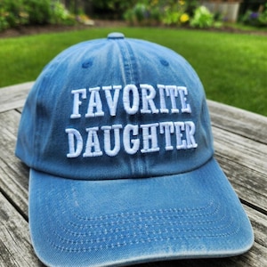 May include: A blue baseball cap with the embroidered text "FAVORITE DAUGHTER" in white. The cap is made of a soft, textured fabric and has a curved brim. The hat is sitting on a wooden surface.