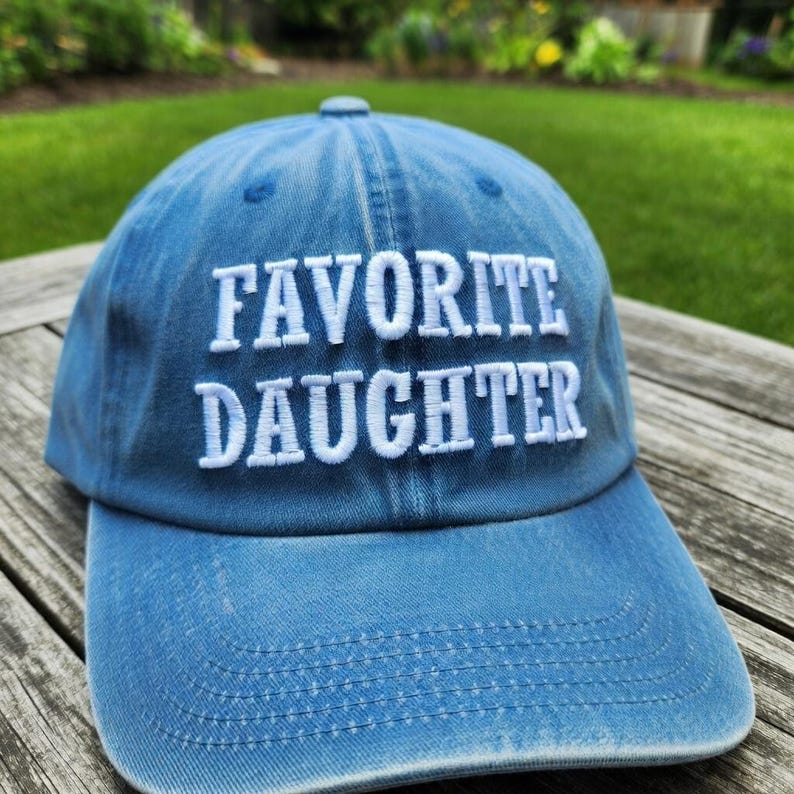 May include: A blue baseball cap with the embroidered text "FAVORITE DAUGHTER" in white. The cap is made of a soft, textured fabric and has a curved brim. The hat is sitting on a wooden surface.