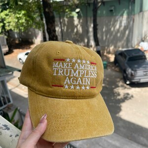 May include: Mustard-colored baseball cap with the text "MAKE AMERICA TRUMPLESS AGAIN" embroidered in white and red. The cap has a curved brim and a button on top. The text is arranged in three lines with stars above and below.