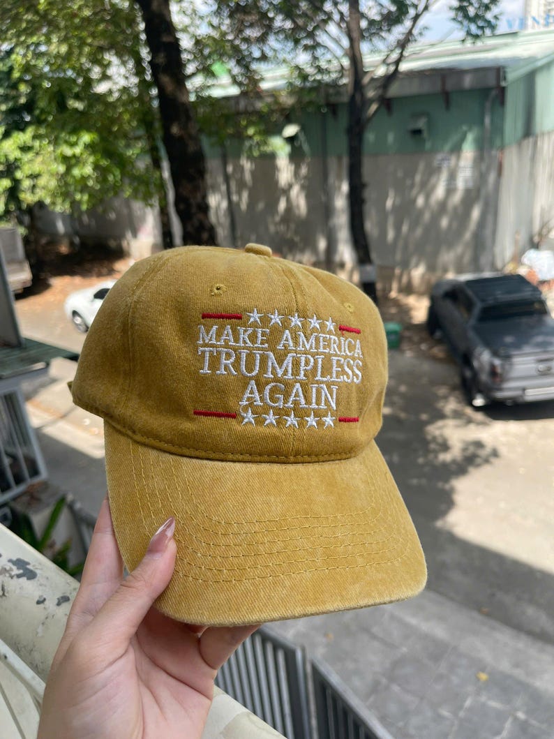 May include: Mustard-colored baseball cap with the text "MAKE AMERICA TRUMPLESS AGAIN" embroidered in white and red. The cap has a curved brim and a button on top. The text is arranged in three lines with stars above and below.
