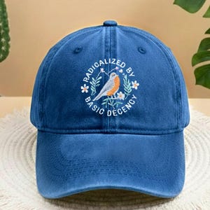 May include: A blue baseball cap featuring the embroidered phrase "RADICALIZED BY BASIC DECENCY" and a bird illustration. The bird is orange, white, and grey, perched on a branch with white flowers. The cap has a curved peak and a button on top.