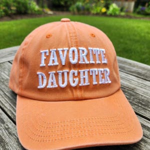 May include: An orange baseball cap with the embroidered text "FAVORITE DAUGHTER" in white. The cap has a curved brim and a fabric strap closure. The hat is made of a soft, durable material.