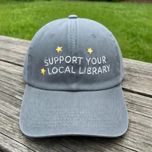 May include: A gray baseball cap with the embroidered text "SUPPORT YOUR LOCAL LIBRARY" in white. The text is accompanied by two yellow star designs. The cap is made of a soft material and has a curved brim.