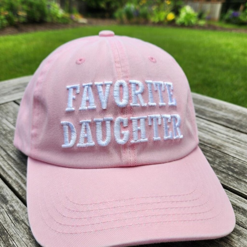 May include: A light pink baseball cap with the embroidered text "FAVORITE DAUGHTER" in white. The cap has a curved brim and a fabric strap closure. The hat is made of a soft, durable material.