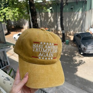 May include: Mustard yellow baseball cap with the text "MAKE AMERICA TRUMPLESS AGAIN" embroidered in white and red. The cap has a curved brim and a six-panel design. A casual accessory.