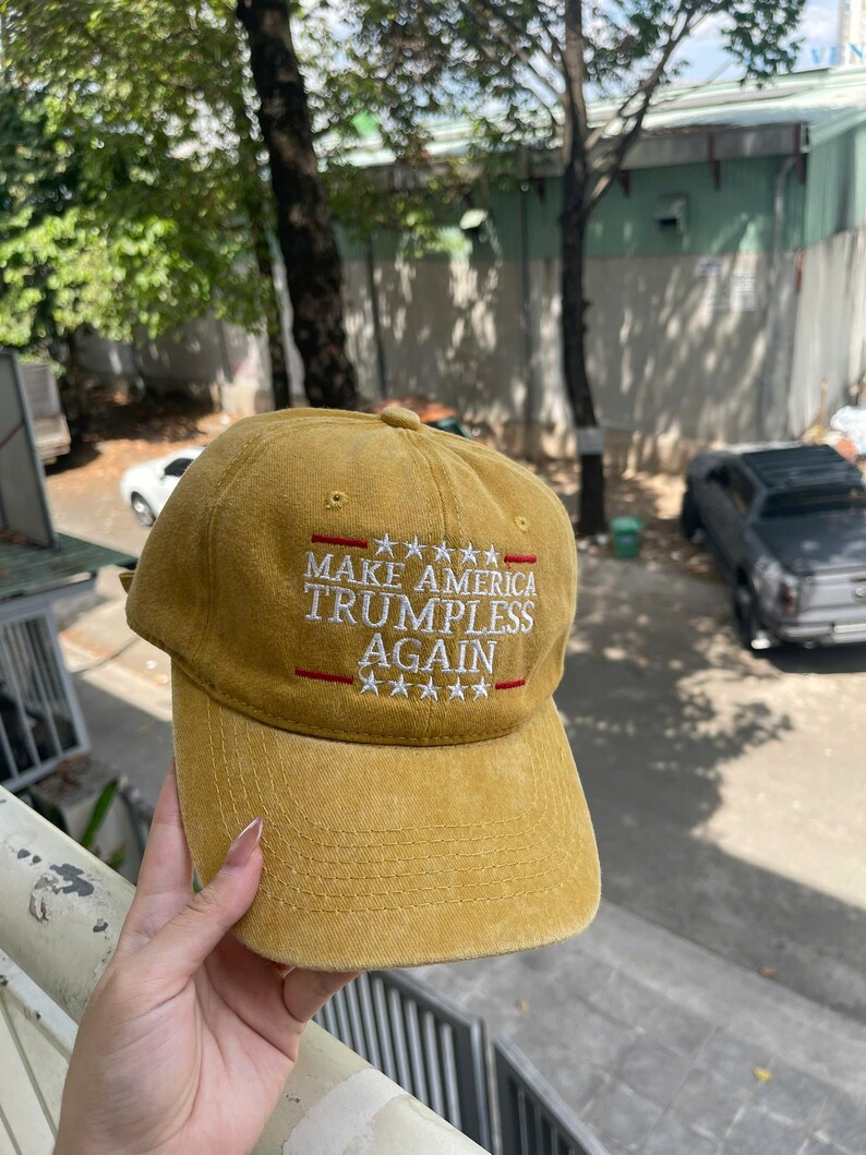 May include: Mustard yellow baseball cap with the text "MAKE AMERICA TRUMPLESS AGAIN" embroidered in white and red. The cap has a curved brim and a six-panel design. A casual accessory.