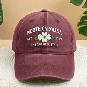 May include: A maroon baseball cap featuring white embroidered text reading "NORTH CAROLINA", a white flower, and the words "EST. 1789 THE TAR HEEL STATE". The cap has a curved peak and a button on top.