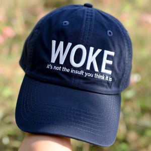 May include: Navy blue baseball cap with the word "WOKE" embroidered in white. Below the word is the phrase "it's not the insult you think it is". The cap has a curved brim and a button on top.