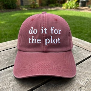 May include: A maroon baseball cap with the phrase "do it for the plot" embroidered in white. The cap is made of a soft fabric and has a curved brim. The hat is sitting on a wooden surface.