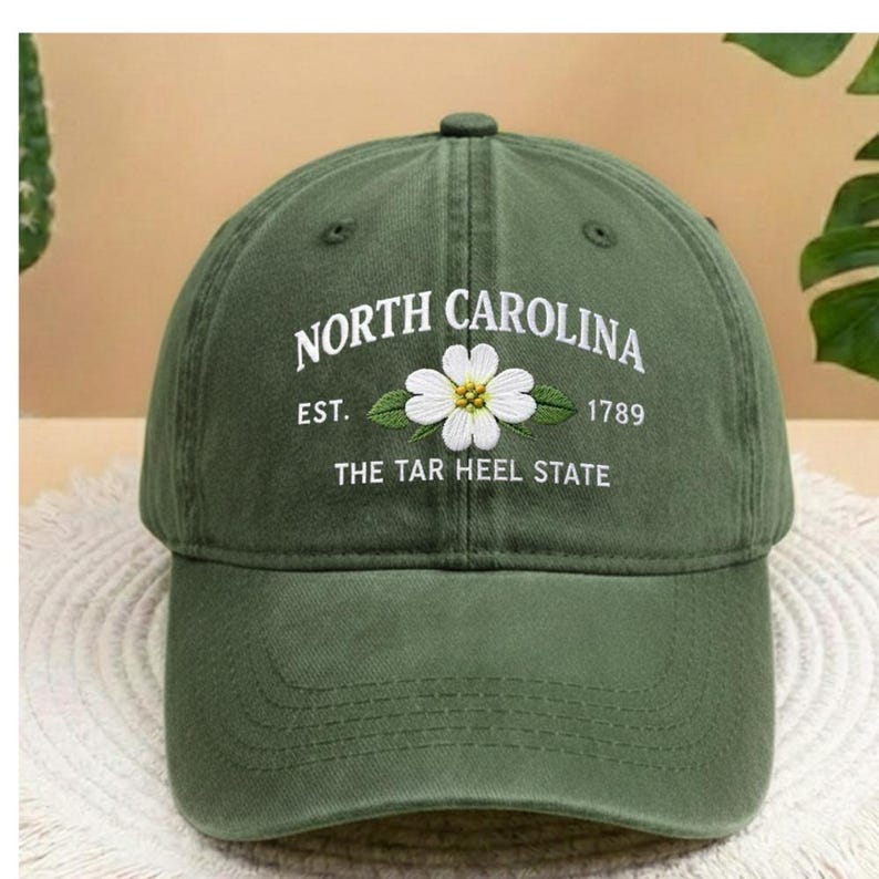 May include: Dark green baseball cap featuring the embroidered words "NORTH CAROLINA EST. 1789 THE TAR HEEL STATE". A white flower with a yellow centre is embroidered below the text. The cap has a curved peak and a button.