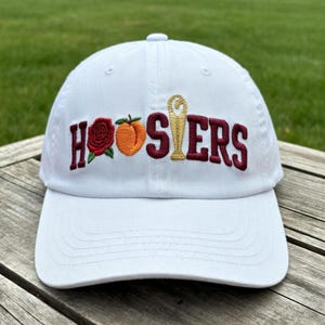 May include: White baseball cap with embroidered text "HOOSIERS" in maroon. The design includes a rose, a peach, and a trophy. The cap has a curved brim and a button on top. The background is green grass.