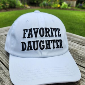 May include: A white baseball cap with the embroidered text "FAVORITE DAUGHTER" in black. The cap has a curved brim and a rounded top, perfect for sun protection. The hat is made of a soft, durable material.