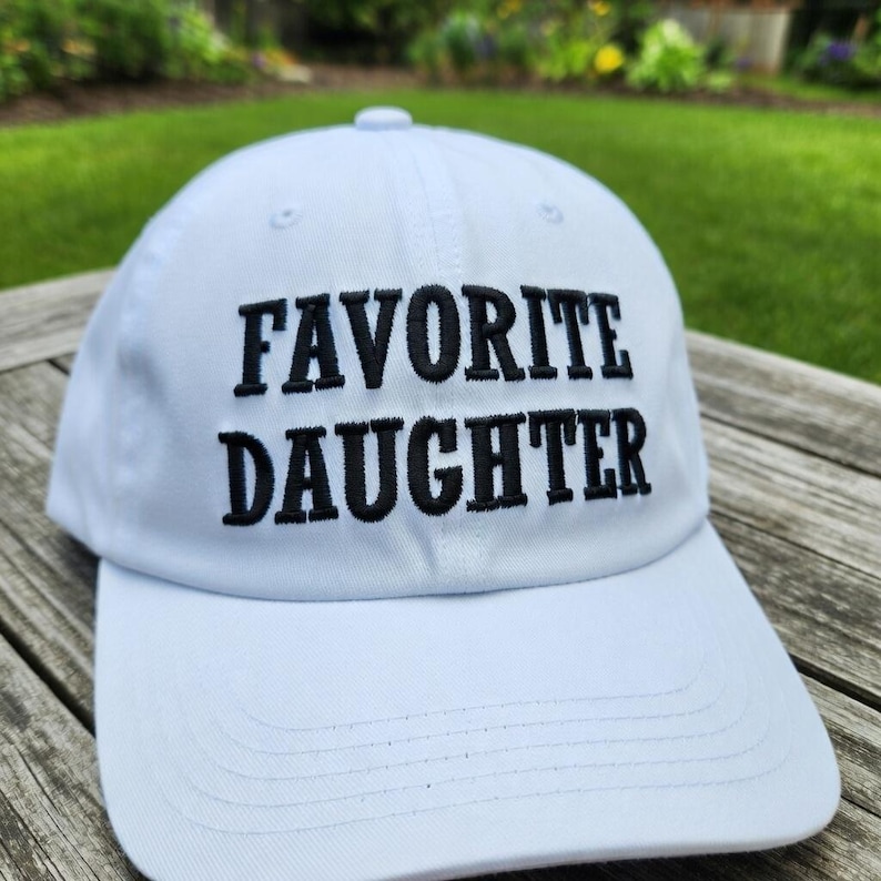 May include: A white baseball cap with the embroidered text "FAVORITE DAUGHTER" in black. The cap has a curved brim and a rounded top, perfect for sun protection. The hat is made of a soft, durable material.