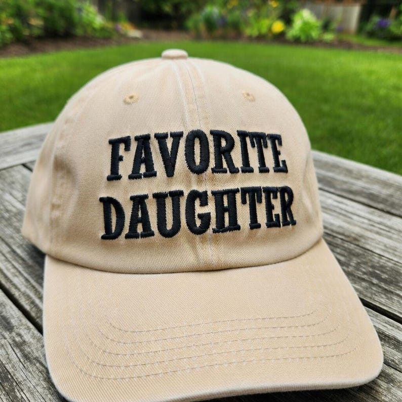 May include: A tan baseball cap with the embroidered text "FAVORITE DAUGHTER" in black. The cap has a curved brim and a fabric strap closure. The background shows a wooden surface and green foliage.