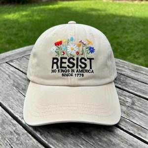 May include: A beige baseball cap with the word "RESIST" embroidered in black, along with the text "NO KINGS IN AMERICA SINCE 1776." The cap features a colorful floral design above the word "RESIST".