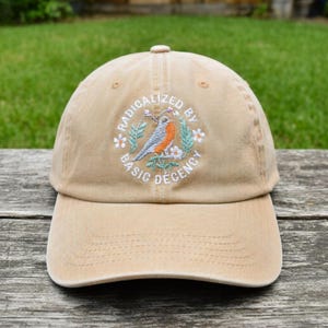 May include: A beige baseball cap with a curved peak. The cap has an embroidered design of a bird, flowers, and the words "RADICALIZED BY BASIC DECENCY" in a circular pattern. The cap is made of a soft fabric and has a button on top.