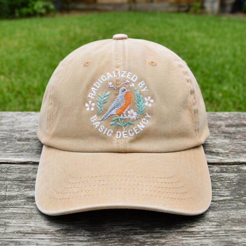 May include: A beige baseball cap with a curved peak. The cap has an embroidered design of a bird, flowers, and the words "RADICALIZED BY BASIC DECENCY" in a circular pattern. The cap is made of a soft fabric and has a button on top.