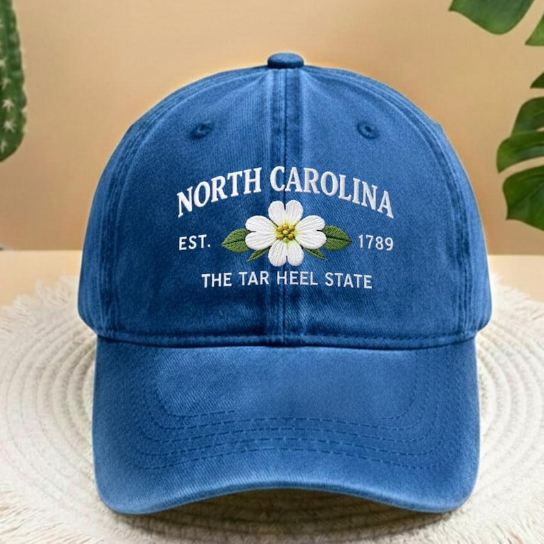 May include: A blue baseball cap featuring the text "NORTH CAROLINA" in white. Below is a white flower with green leaves, and the text "EST. 1789 THE TAR HEEL STATE". The cap has a curved peak and a button on top.