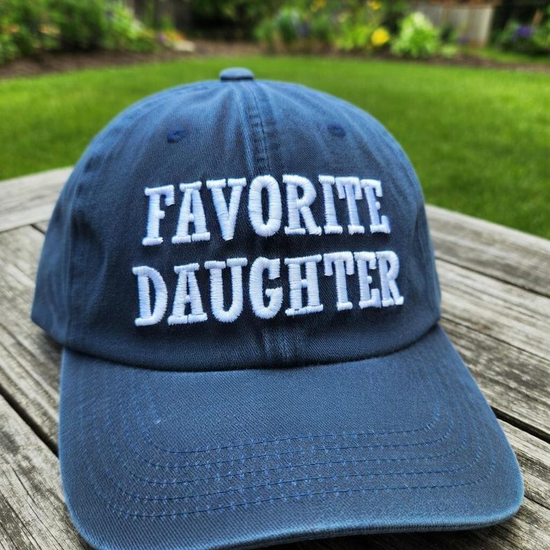 May include: A navy blue baseball cap with the embroidered text "FAVORITE DAUGHTER" in white. The cap is made of a soft fabric and has a curved brim. The hat is sitting on a wooden surface.