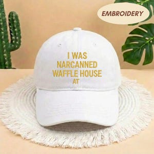 May include: White baseball cap with the embroidered text "I WAS NARCANNED WAFFLE HOUSE AT" in gold. The cap has a curved brim and a rounded crown. The word "EMBROIDERY" is in a tan oval in the upper right corner.