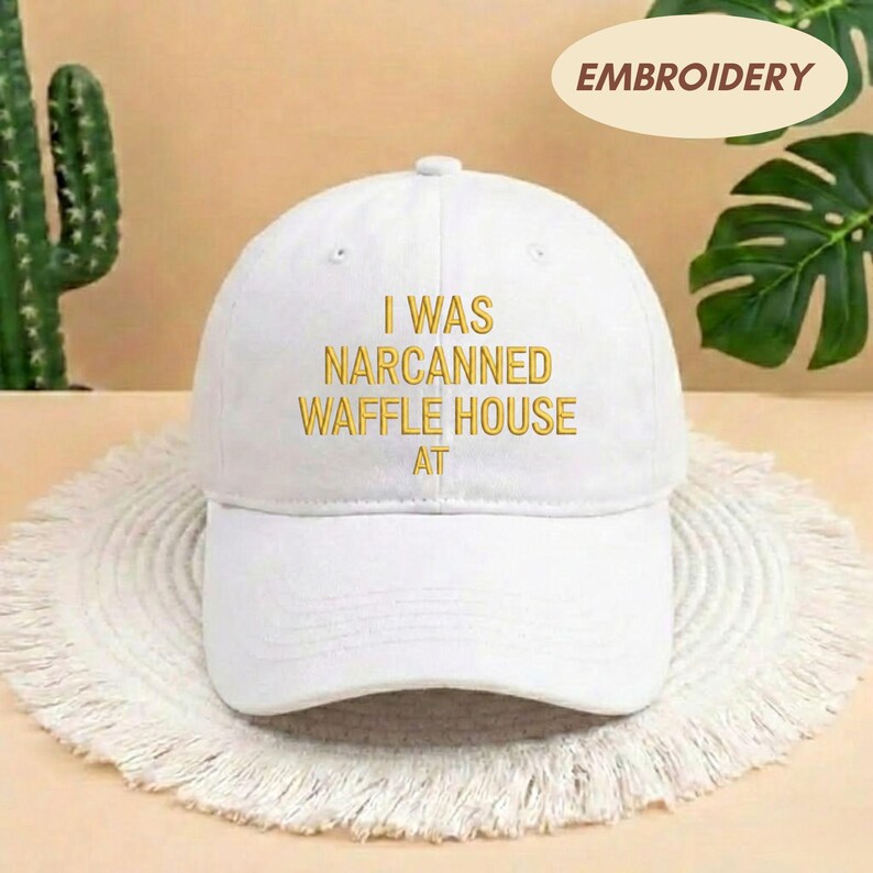 May include: White baseball cap with the embroidered text "I WAS NARCANNED WAFFLE HOUSE AT" in gold. The cap has a curved brim and a rounded crown. The word "EMBROIDERY" is in a tan oval in the upper right corner.