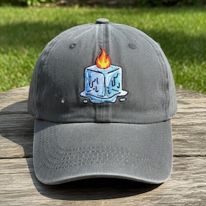 May include: A gray baseball cap with a curved brim. The cap features an embroidered design of a blue ice cube with a flame on top. The ice cube is melting. The cap is made of a soft material.