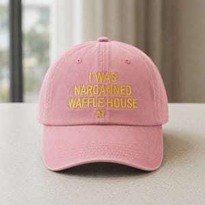 May include: A pink baseball cap with the embroidered text "I WAS NARCANNED WAFFLE HOUSE AT" in gold thread. The cap has a curved brim and a button on top.