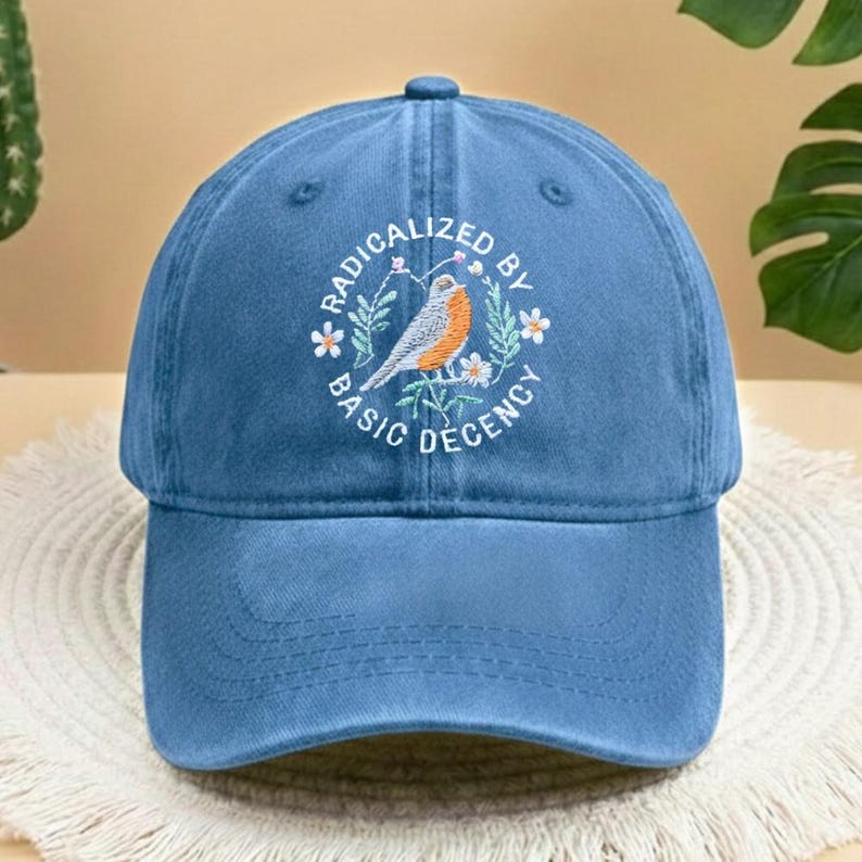 May include: A blue baseball cap with a curved peak. The cap has an embroidered design of a bird, flowers, and the text "RADICALIZED BY BASIC DECENCY" in a circular pattern. The hat is made of a soft, textured fabric.