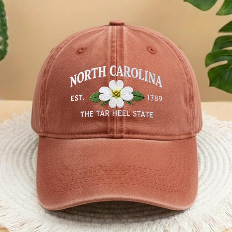 May include: A terracotta baseball cap featuring embroidered text and a floral design. The text reads "NORTH CAROLINA" above "EST. 1789" and "THE TAR HEEL STATE". The flower is white with a yellow centre and green leaves.