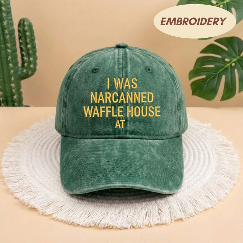 May include: Green baseball cap with the text "I WAS NARCANNED WAFFLE HOUSE AT" in yellow. The cap is made of a worn-looking fabric and has a curved brim. The word "EMBROIDERY" is in a tan oval in the upper right corner.