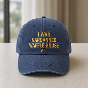 May include: A navy blue baseball cap with the embroidered text "I WAS NARCANNNED WAFFLE HOUSE AT" in gold thread. The cap has a curved brim and a classic six-panel design, suitable for casual wear.
