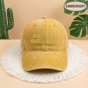 May include: Mustard yellow baseball cap with the embroidered text "I WAS NARCANNNED WAFFLE HOUSE AT". The cap is sitting on a white, fringed placemat. The word "EMBROIDERY" is in a green oval in the upper right corner.