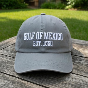 May include: A gray baseball cap with the text "GULF OF MEXICO EST. 1550" embroidered in white. The cap is made of a soft fabric and has a curved brim. The cap is sitting on a wooden surface.