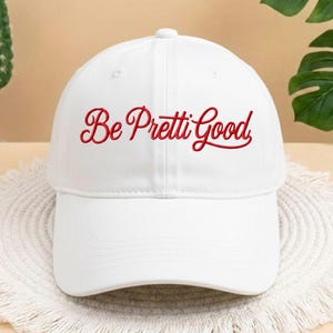 May include: A white baseball cap with the embroidered phrase "Be Pretti Good" in red script across the front. The cap has a curved brim and a button on top. The hat is sitting on a white, woven surface.