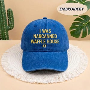 May include: A blue baseball cap with the embroidered text "I WAS NARCANNED WAFFLE HOUSE AT" in yellow. The cap has a curved brim and a fabric strap closure. The word "EMBROIDERY" is printed on a white oval in the upper right corner.