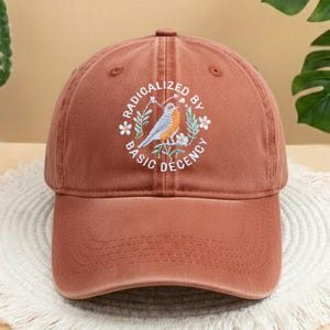 May include: A terracotta-coloured baseball cap with a curved peak. The cap has an embroidered design of a bird, flowers, and the text "RADICALIZED BY BASIC DECENCY" in a circular pattern. The cap is made of a soft, textured fabric.
