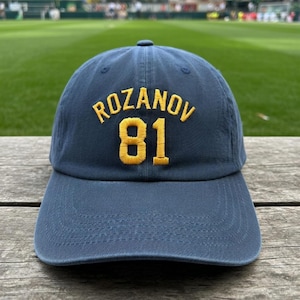 May include: Navy blue baseball cap with the name "ROZANOV" and the number "81" embroidered in yellow. The cap has a curved brim and a classic six-panel design. The background shows a green field.