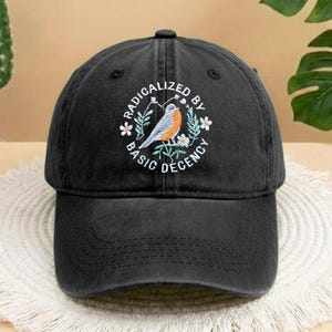 May include: Black baseball cap with a curved peak. The cap has an embroidered design of a bird, flowers, and the text "RADICALIZED BY BASIC DECENCY" in a circular pattern. The cap is made of a soft, textured fabric.