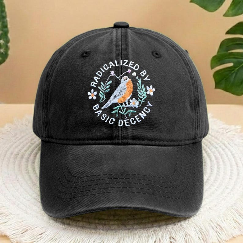 May include: Black baseball cap with a curved peak. The cap has an embroidered design of a bird, flowers, and the text "RADICALIZED BY BASIC DECENCY" in a circular pattern. The cap is made of a soft, textured fabric.