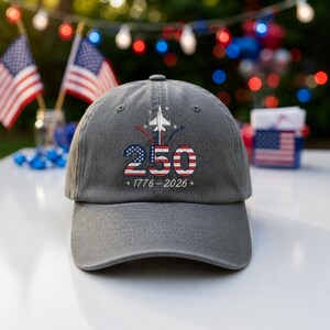 May include: A charcoal gray baseball cap featuring an embroidered design with the number 250 in red, white, and blue, a white fighter jet silhouette, and the text 1776 - 2026. The cap has a curved brim and a structured crown with eyelets.