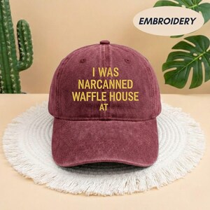 May include: A maroon baseball cap with the embroidered text "I WAS NARCANNED WAFFLE HOUSE AT" in yellow. The hat is displayed on a white, fringed placemat. The word "EMBROIDERY" is printed on a white oval.